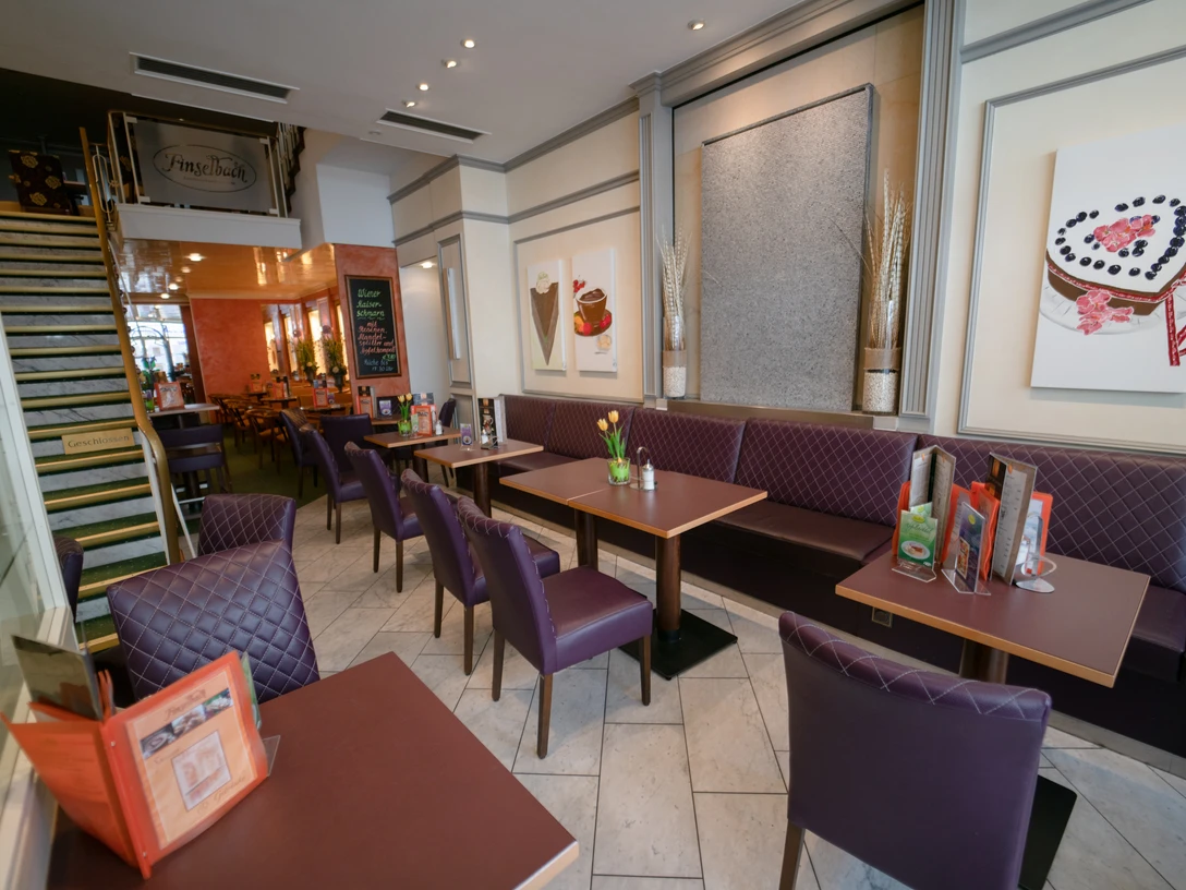 Innenansicht Das Bild zeigt die stillvolle Innenansicht des Cafés mit dunklen Holztischen, gepolsterten Sitzbänken und moderner Wanddekoration. Eine Treppe führt nach oben, während im Hintergrund ein weiterer Sitzbereich mit warmen Licht zu sehen ist.