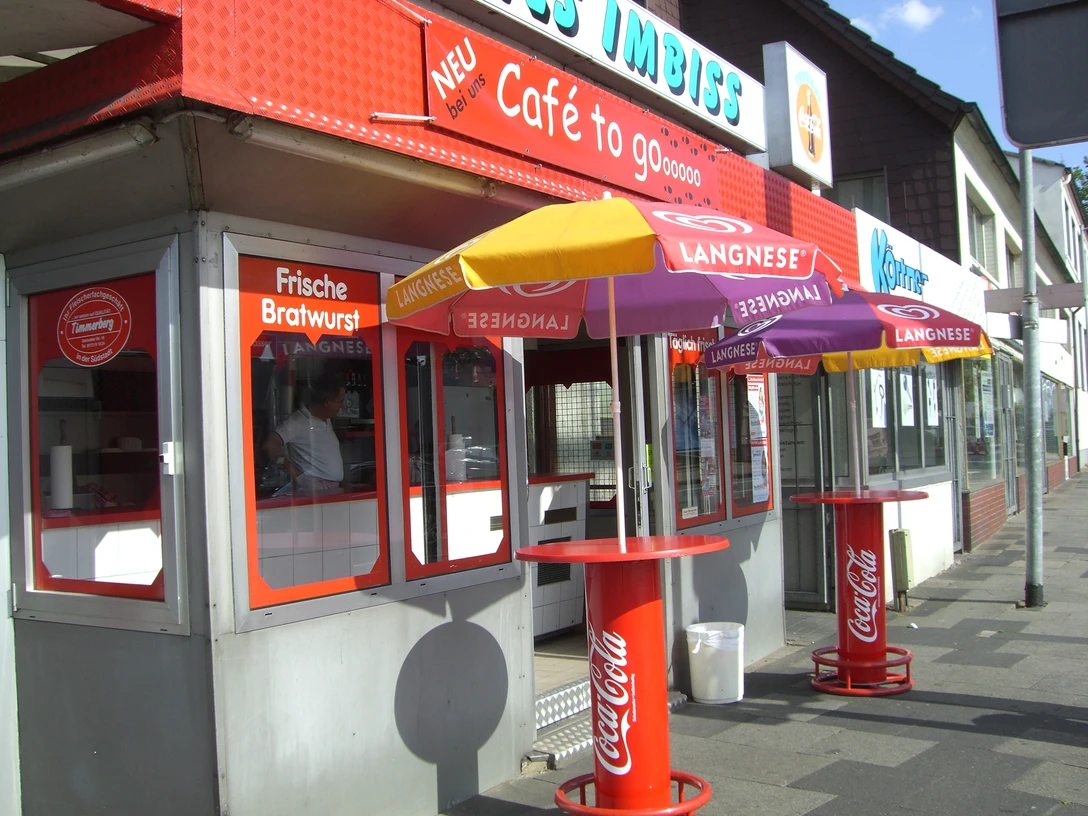 Außenansicht Auf dem Bild ist die Außenansicht des Imbisses zu sehen, dessen Fassade in einem ansprechenden Rot und Grau gestaltet ist. Vor dem Imbiss stehen einladende Sonnenschirme, die den Gästen Schatten spenden, während sie ihre köstliche Currywurst genießen.