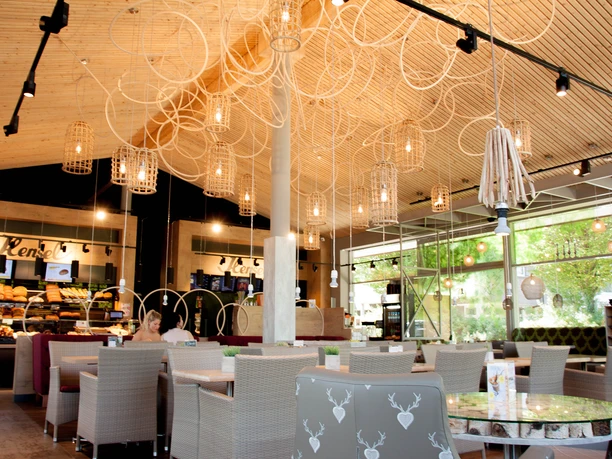 Innenansicht Das Bild zeigt die Innenansicht der Bäckerei mit einer stillvollen Deckenbeleuchtung aus mehreren hängenden Lampen. Im Hintergrund sind eine große Fensterfront mit Blick ins Grüne sowie eine Theke mit verschiedenem Gebäck zu sehen.