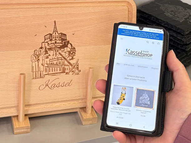 Mein KasselShop Auf einem Handy wird der Mein KasselShop gezeigt. Im Hintergrund befinden sich Kassel Souvenirs.