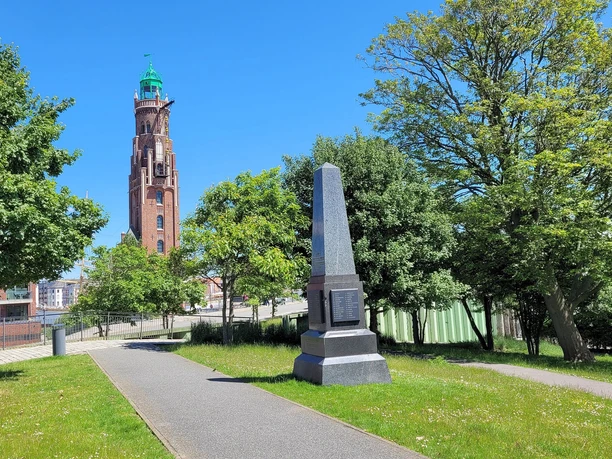 20220603_141407 (c) Tanja Mehl_Erlebnis Bremerhaven_WEB.jpg Obelisk zur Erinnerung der auf See gebliebenen Menschen und dahinter die Gedenkdalben mit Namensschildern der auf See bestatteten Menschen mit Blick auf den Simon-Loschen-Leuchtturm