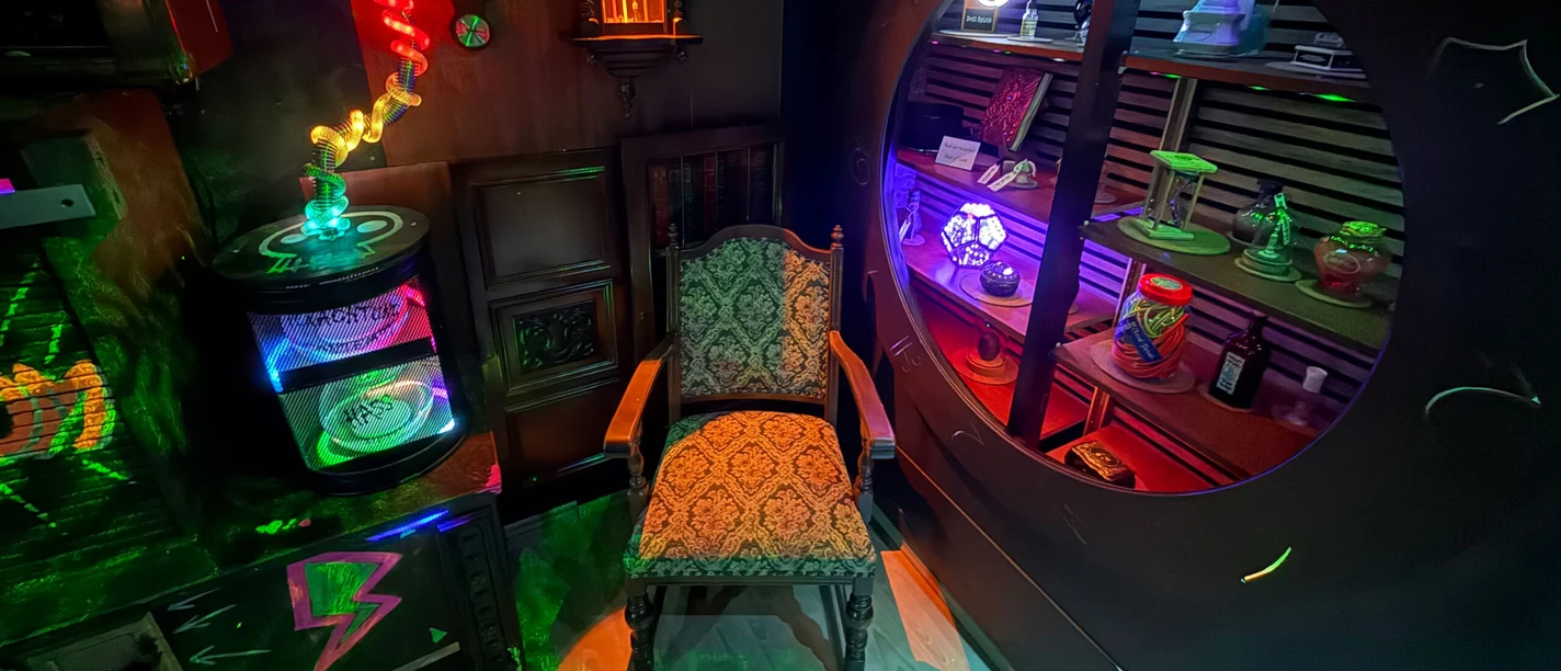 Escape Room MAGIC BOOM Colorful "Magic School" escape room featuring glowing objects, a shelf, and an old chair.