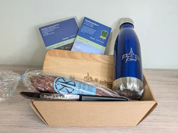 Kassel Geschenkkorb Ein Geschenkkorb mit Souvenirartikeln von Kassel. Es befindet sich eine Trinkflasche mit Herkulesmotiv, ein Schneidemesser und Schneidebrettchen mit Kassel Skyline, eine Ahle Wurscht und Kartenmaterial in dem Geschenkkorb.