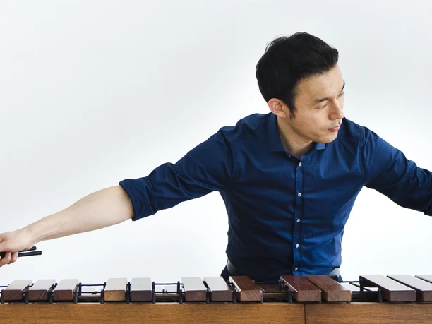 Fumito Nunoya – Meister der Marimba Fumito Nunoya spielt mit vier Schlägeln auf einem großen Marimbaphon, gekleidet in einem blauen Hemd.