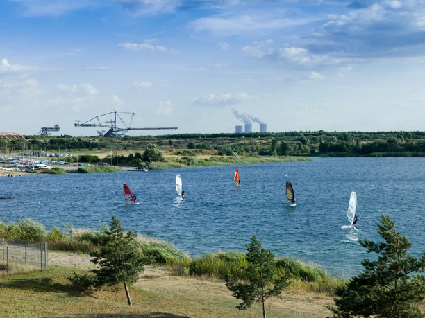 Auf dem Markkleeberger See, der von Natur umgeben ist, sind zahlreiche Windsurfer unterwegs, im Hintergrund sind der Bergbau-Technik-Park und das Kraftwerk Lippendorf zu sehen
