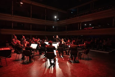 Silvesterkonzerte Kammerorchester mit Dirigent auf rot beleuchteter Bühne vor Publikum.