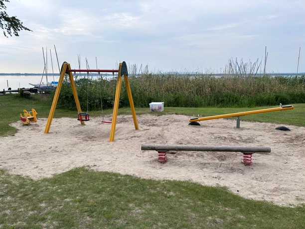 A playground with a few pieces of play equipment on a sandy area in front of Lake Steinhude. In the background, you can see some reeds. The playground has a seesaw, a swing, and rocking animals.