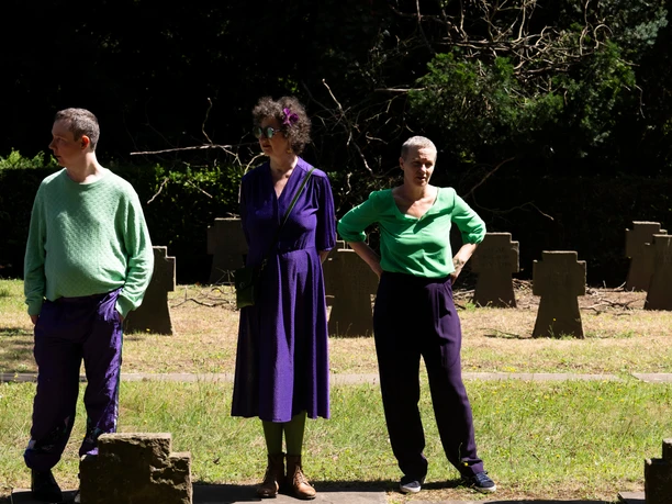 BOTIN Three people dressed in green and purple are standing among gravestones in the sunlight.