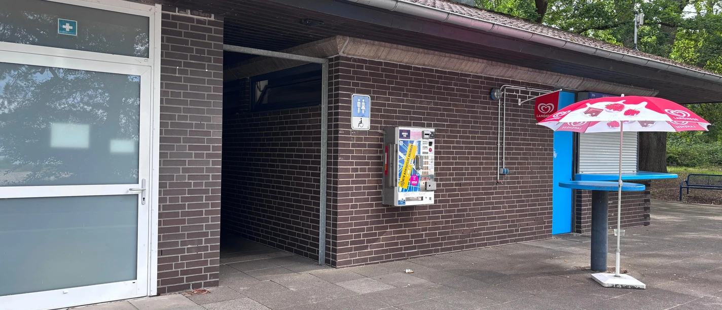 Toilette Kiosk am Badestrand Ein braunes Steingebäude mit Sonnenschirm davor und einem Tor mit dem Eingang zur WC-Anlage.