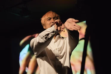 Wang Chong An actor with blond hair, wearing a white shirt, is gesturing on a dark theater stage