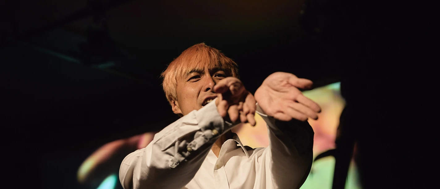 Wang Chong An actor with blond hair, wearing a white shirt, is gesturing on a dark theater stage