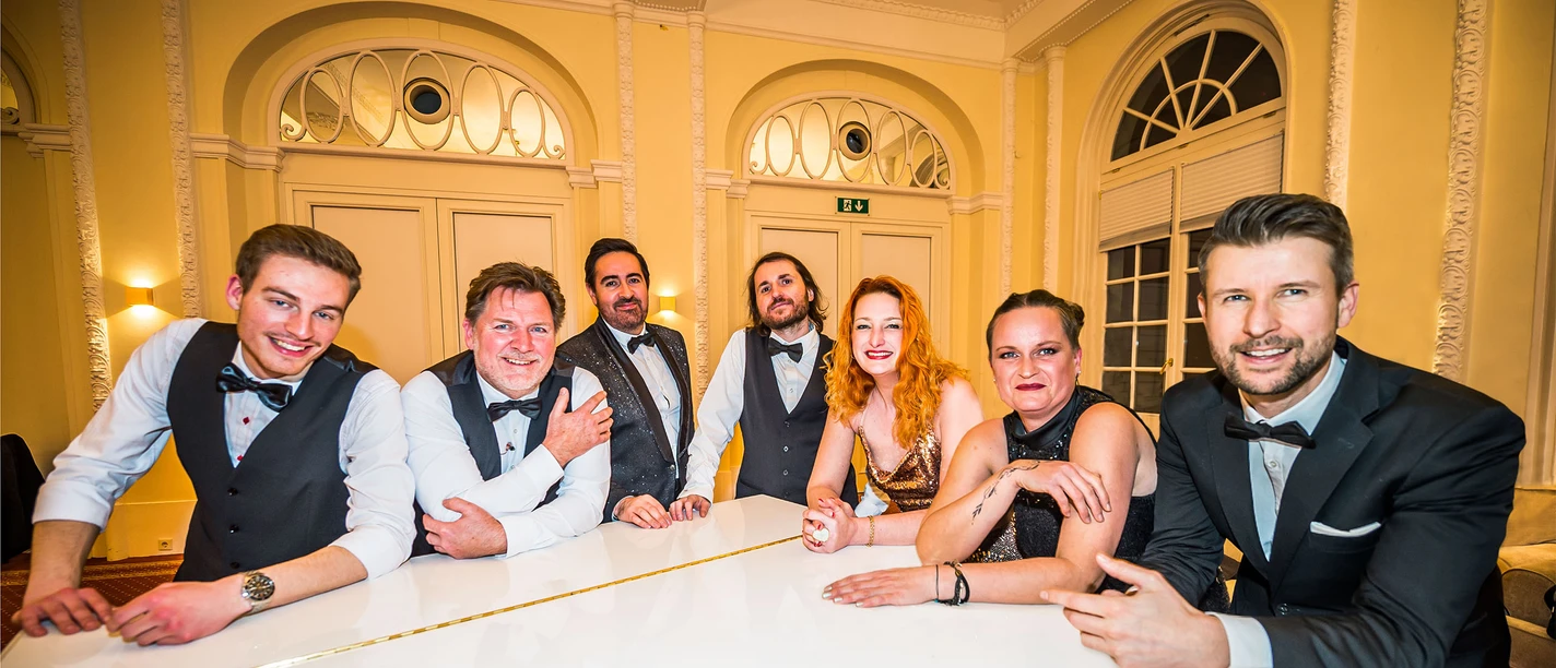 Casino Royale bei der Büsumer Sommerparty auf der Watt-Tribüne Eine Gruppe von 7 Personen stehen um einen weißen Flügel herum.