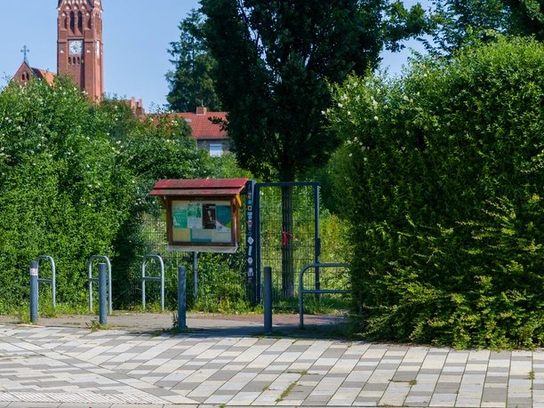 Zolli T0473 (c) Helmut Gross_Erlebnis Bremerhaven_WEB.jpg Eingang zum Park Zolli mit Infokasten vor grünen Büschen. Im Hintergrund der Kirchturm der Pauluskirche.