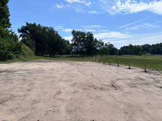 P1 (1) Parkfläche auf Sandboden mit Trennzaun neben einer Wiese. Im Hintergrund sind Bäume unter blauem Himmel zu sehen.