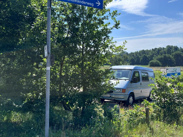 P1 (1).png Parkfläche auf Sandboden mit Trennzaun neben einer Wiese. Im Hintergrund sind Bäume unter blauem Himmel zu sehen.