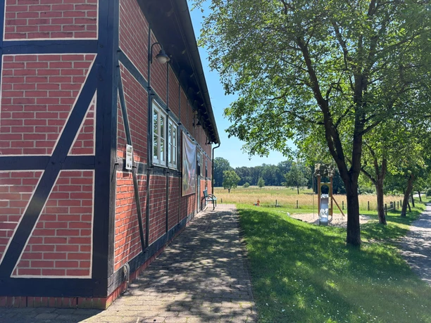 Toilette Haus des Gastes - außen Ein gepflasterter Weg führt rechts am Fachwerkhaus zur Toilettentür, rechts davon geht ein Rasenhang abwärts und im Hintergrund ist ein Spielplatz zu sehen.