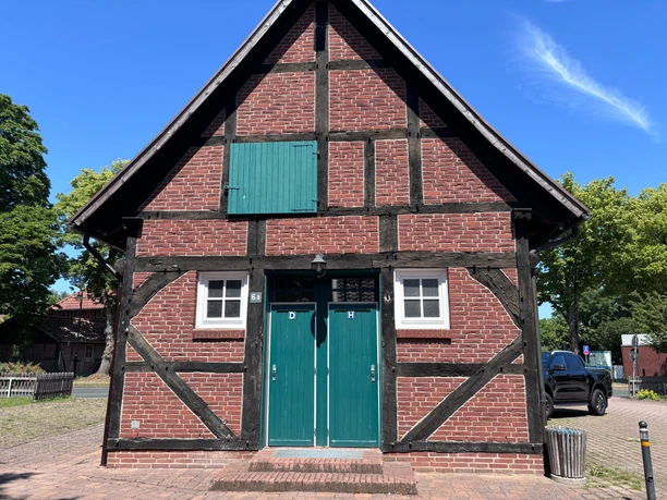 Toilette Aloys-Bunge-Platz (2) Blick auf ein altes Fachwerkhaus mit grüner Tür und jeweils einem kleinen Fenster daneben. Oben am Haus ist eine grüne Fensteröffnung.
