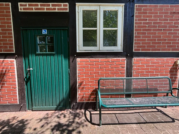 Toilette Haus des Gastes - außen Blick auf den Seiteneingang eines Fachwerkhauses (Haus des Gastes). Eine grüne Bank steht neben einer Tür zur Behindertentoilette.