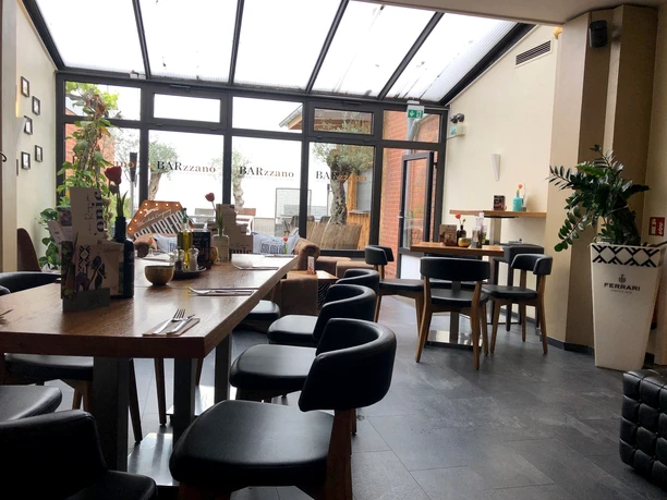 Barzzano Nienburg Ein helles Restaurant mit modernem Interieur, Holztischen und schwarzen Stühlen in einem Wintergarten.