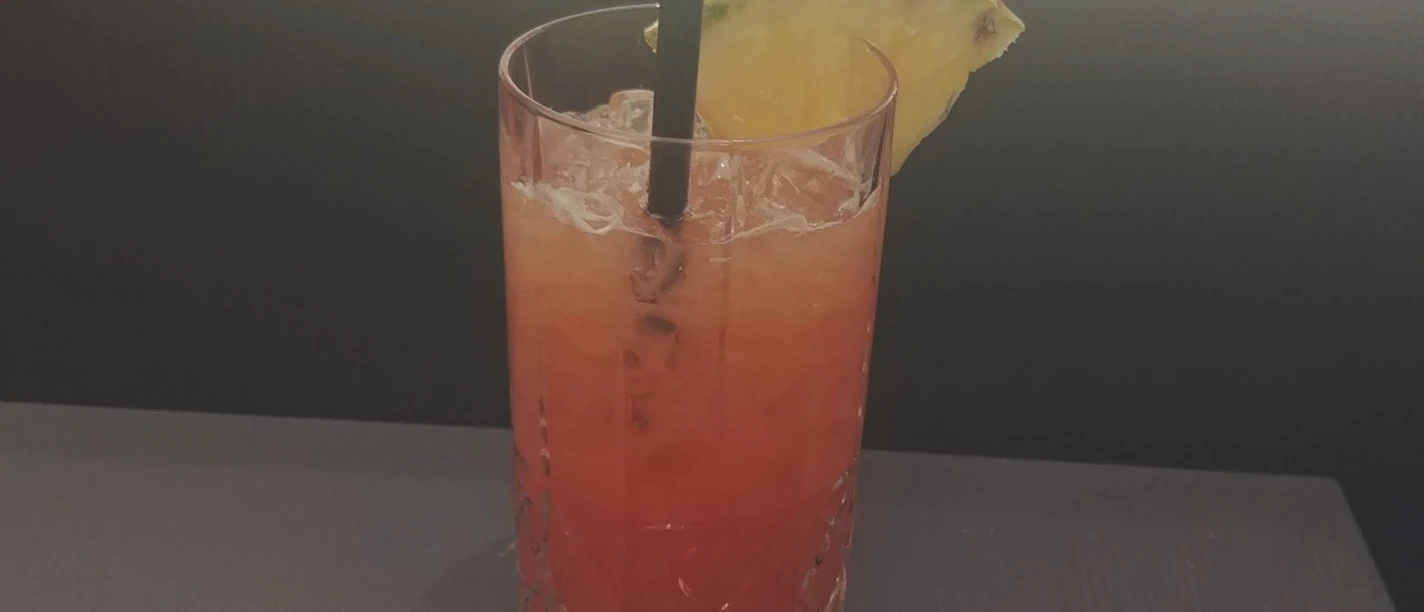 Een verfrissende rode cocktail in een hoog glas met een schijf ananas en een rietje bovenop.