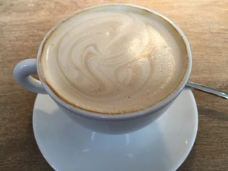 Koffiekopje met melkschuim op een houten tafel, vergezeld van een theelepeltje.