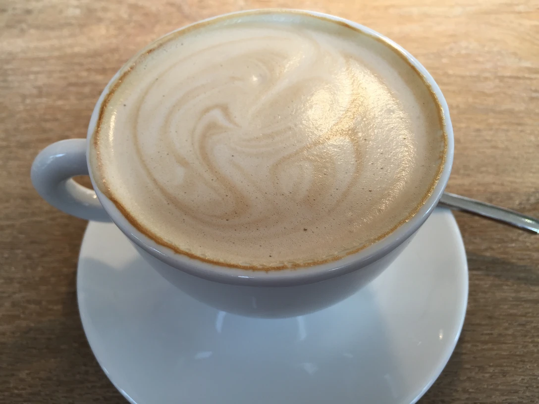 Kaffeetasse mit schaumiger Milchoberfläche auf Holztisch, begleitet von einem Teelöffel.