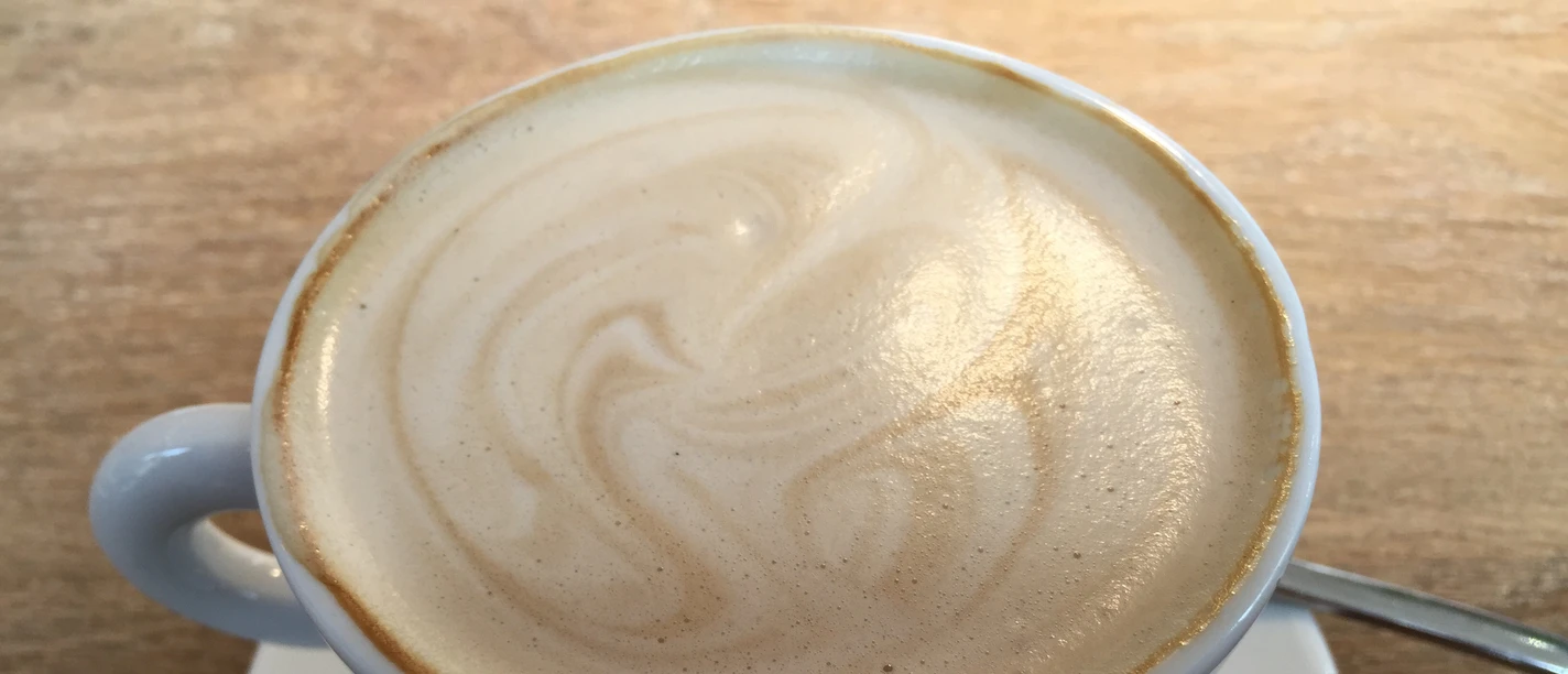 Coffee cup with frothy milk surface on a wooden table, accompanied by a teaspoon.
