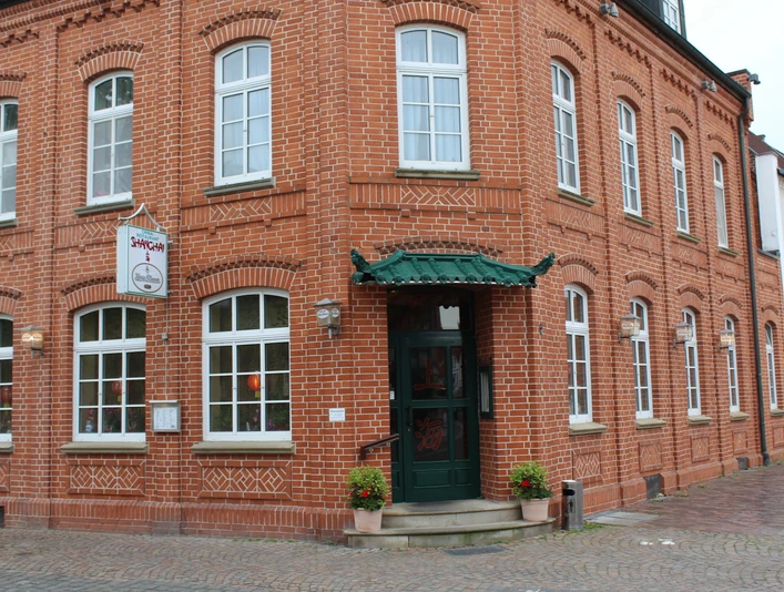 Rotes Backsteingebäude mit grünem Eingang und mehreren Fenstern, gelegene an einem gepflasterten Gehweg.