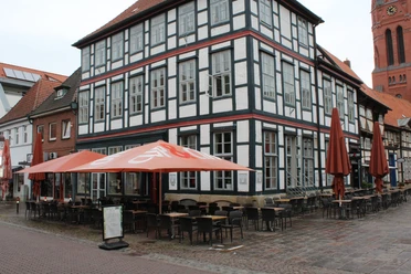 Vakwerkhuis met caféterras, stoelen en tafels buiten, onder een blauwe hemel en rode parasols.