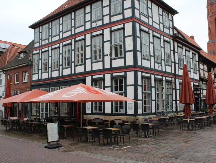 Cup & Cino Fachwerkhaus mit Café-Terrasse, Stühle und Tische im Freien, bei blauem Himmel und roten Sonnenschirmen.