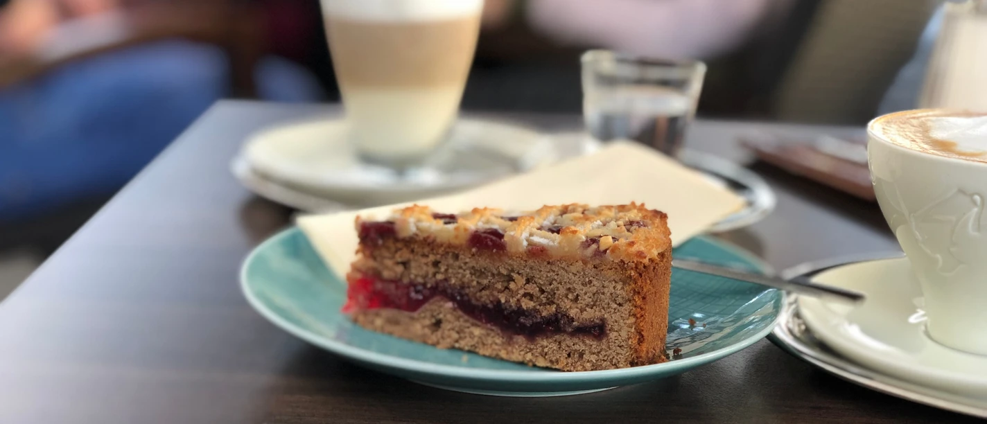 A slice of crumble cake with red filling on a blue plate, a café in the background.