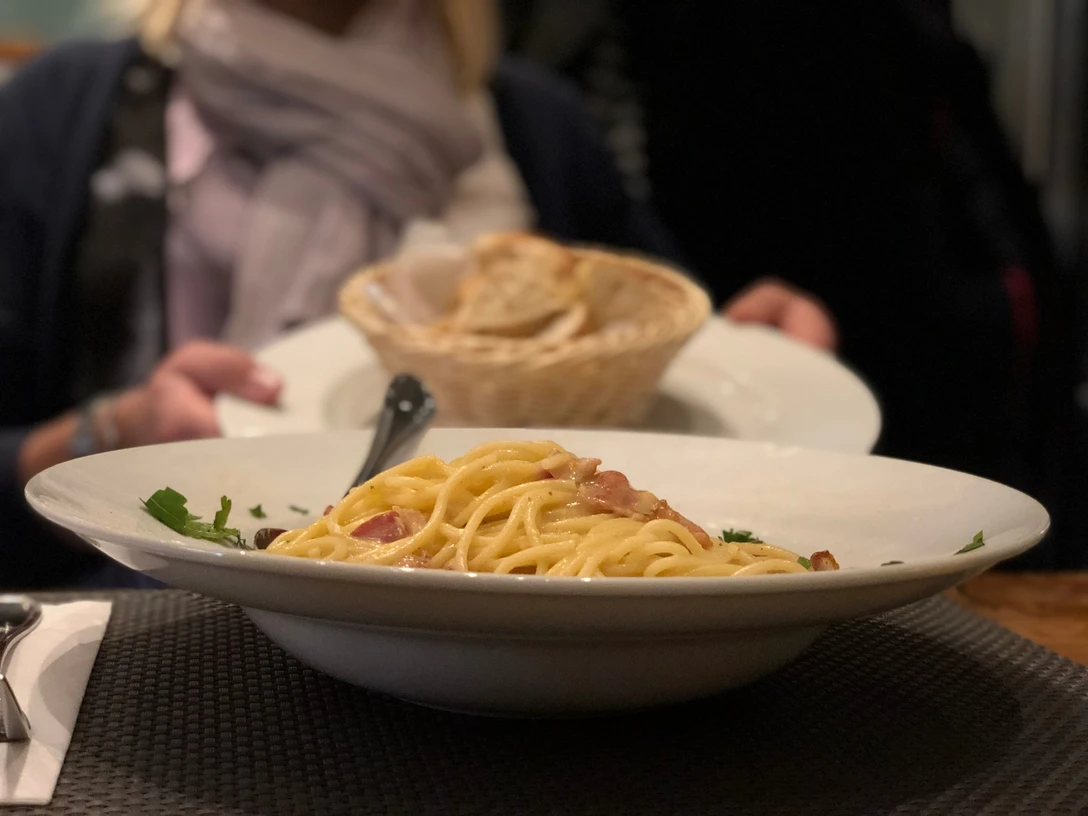 Pasta Ein Teller Spaghetti Carbonara mit Speck und Petersilie, dahinter ein Korb mit Brot auf einem Tisch.