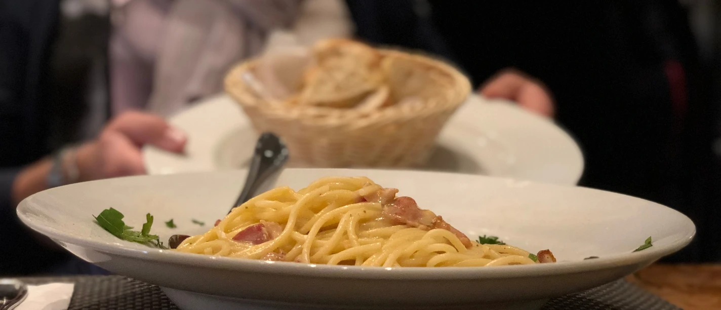 Pasta Ein Teller Spaghetti Carbonara mit Speck und Petersilie, dahinter ein Korb mit Brot auf einem Tisch.