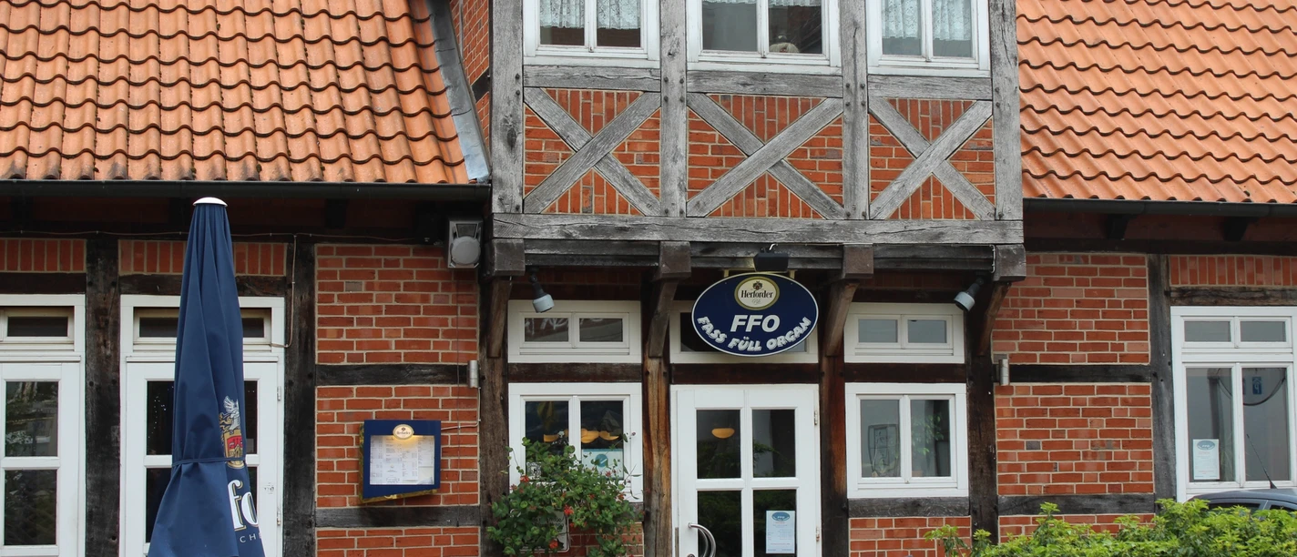 Brick building in half-timbered style with roofed canopy and weather vane, surrounded by plants.