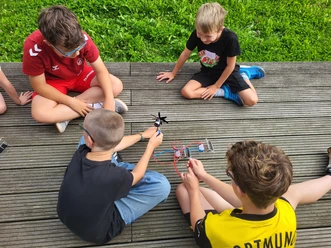 Sommerferien-Programm 2026 Kinder bauen auf einem Holzdeck gemeinsam ein kleines Propeller-Experiment.