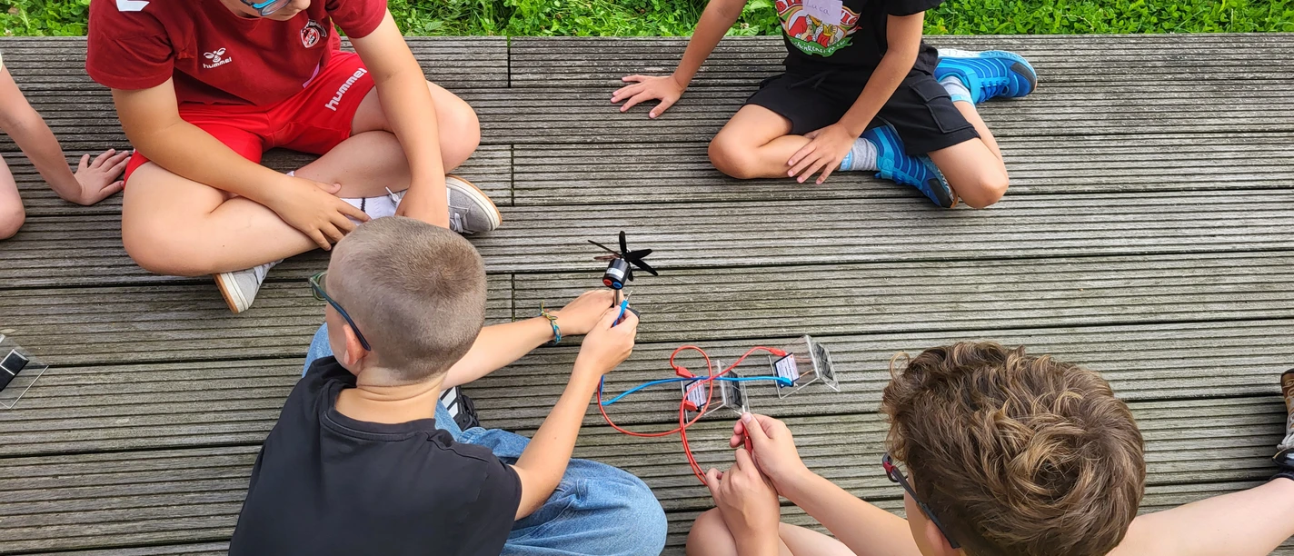 Sommerferien-Programm 2026 Kinder bauen auf einem Holzdeck gemeinsam ein kleines Propeller-Experiment.