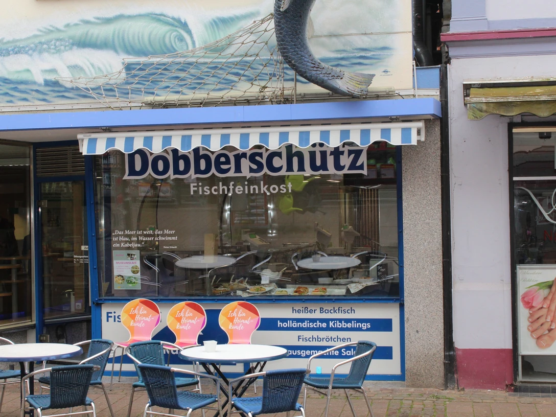 Ein Fischgeschäft mit blauer Markise und einem großen Fisch an der Wand. Davor stehen Tische und Stühle.