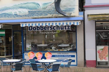 Dobberschütz Ein Fischgeschäft mit blauer Markise und einem großen Fisch an der Wand. Davor stehen Tische und Stühle.