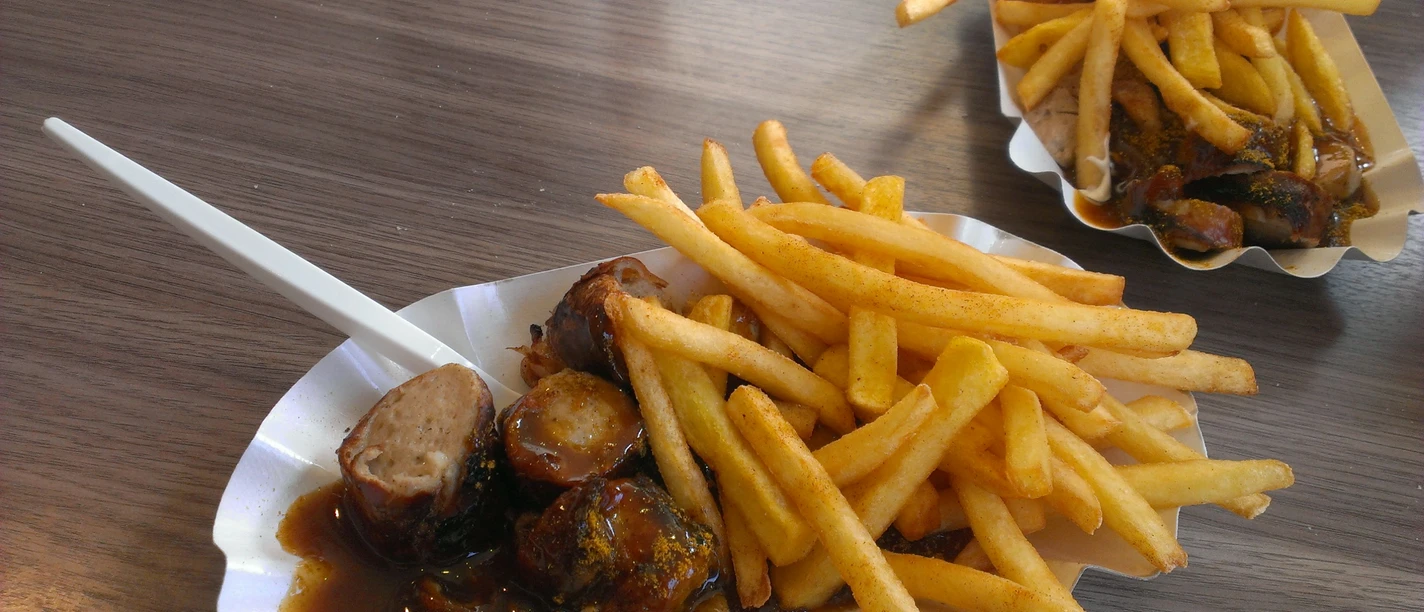 Delicious currywurst with crispy chips, served in a cardboard bowl with a plastic fork.