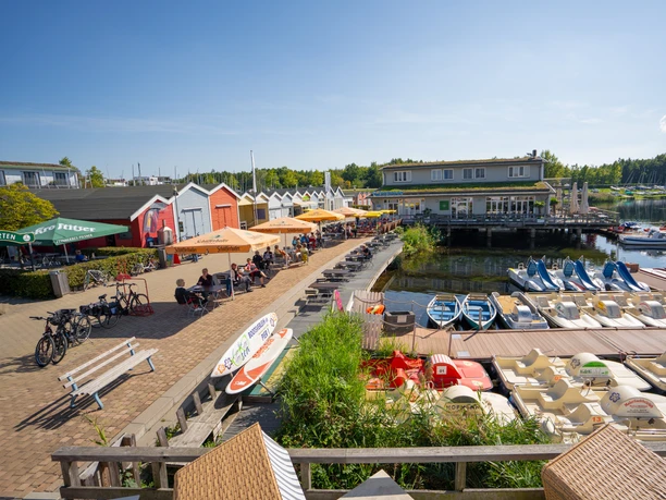 Links befindet sich der Biergarten am Hafen Pier 1 mit zahlreichen Gästen unter Sonnenschirmen, mittig steht ein Gastronomiegebäude auf Stelzen, davor ein Steg mit vielen Tret- und Paddelbooten und in der Ferne am Ufer des Cospudener Sees weitere Segelboote