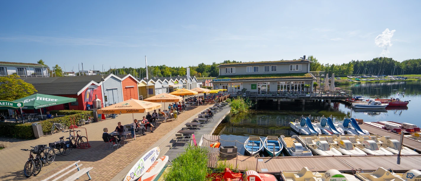 Pier 1 am Cospudener See - Leipziger Neuseenland Links befindet sich der Biergarten am Hafen Pier 1 mit zahlreichen Gästen unter Sonnenschirmen, mittig steht ein Gastronomiegebäude auf Stelzen, davor ein Steg mit vielen Tret- und Paddelbooten und in der Ferne am Ufer des Cospudener Sees weitere Segelboote
