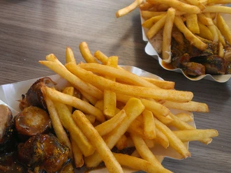 Two cardboard trays filled with crispy chips and pieces of currywurst dipped in sauce on a wooden table.