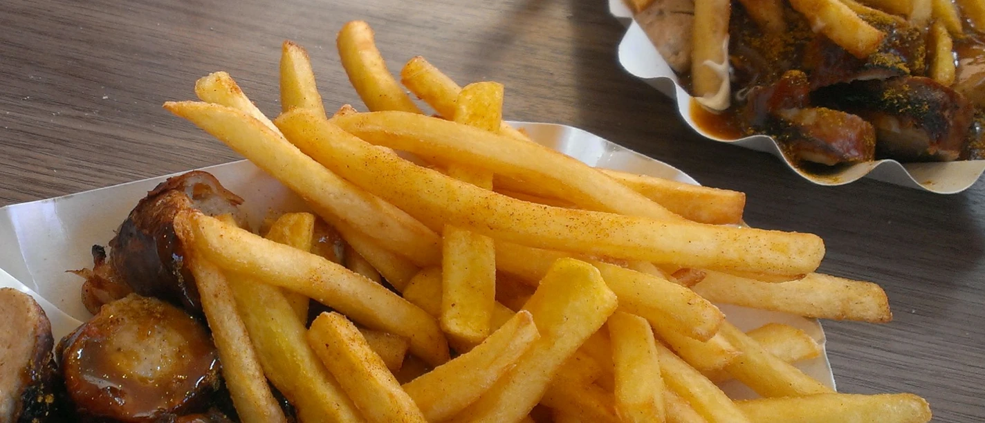 Two cardboard trays filled with crispy chips and pieces of currywurst dipped in sauce on a wooden table.