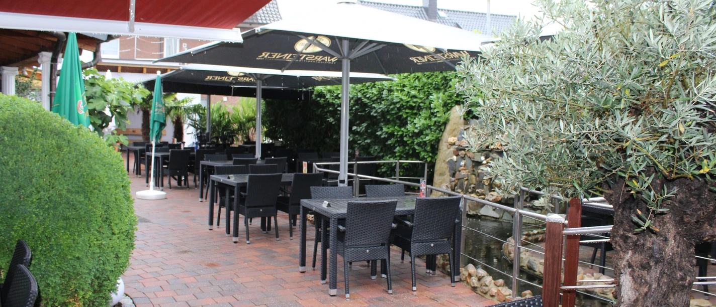 Overdekt buitenterras met zwarte stoelen en tafels, omringd door groene planten.