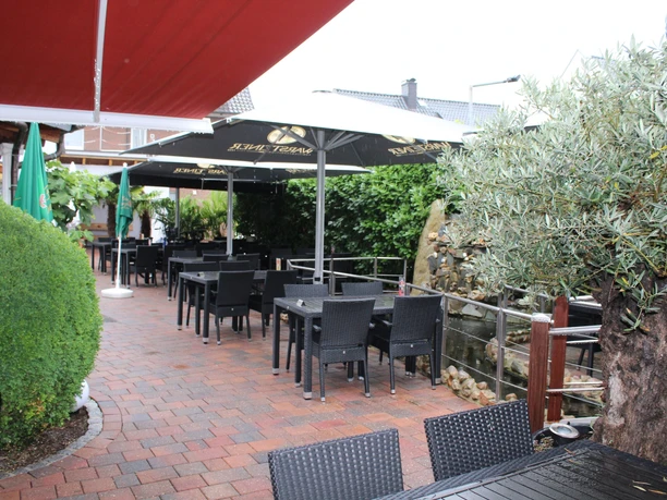 Covered outdoor terrace with black chairs and tables, surrounded by green plants.