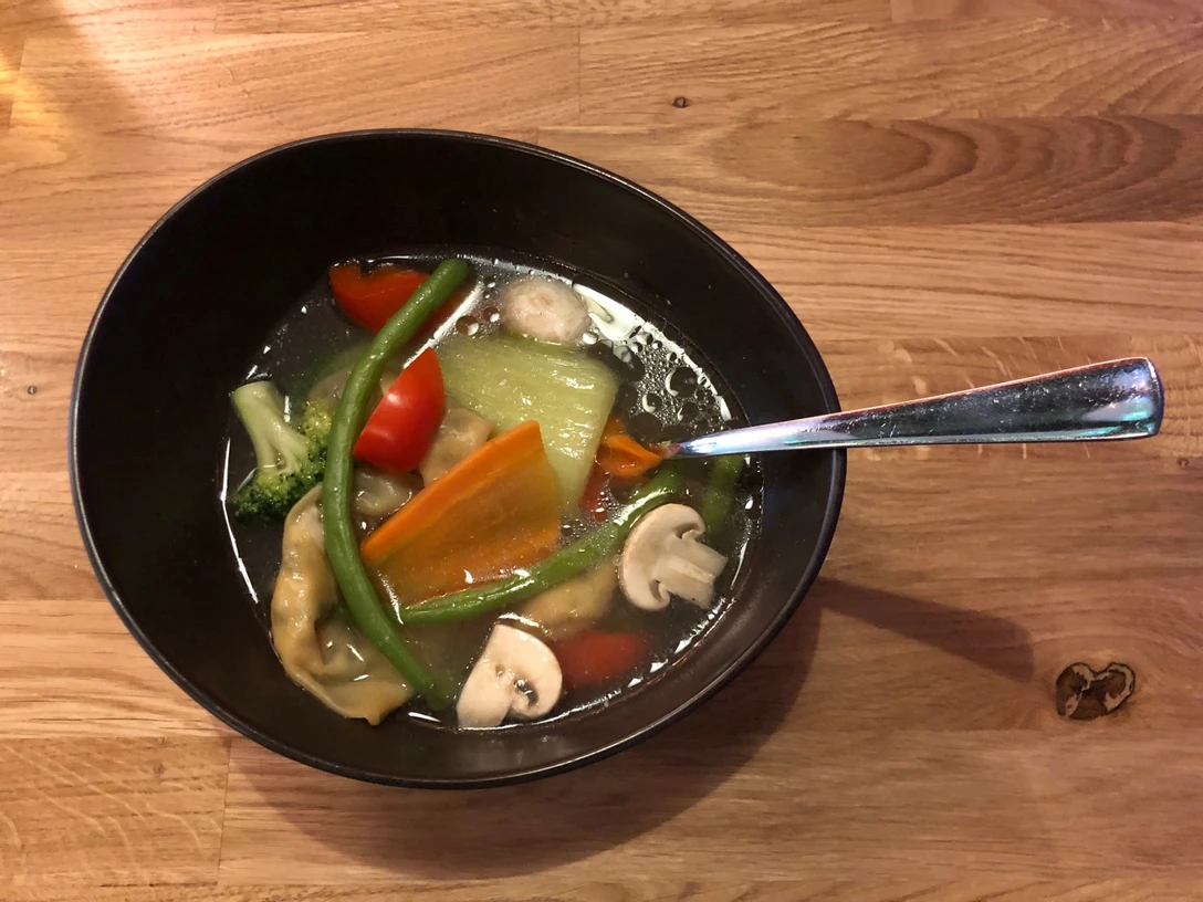 Chinesische Suppe Eine Schüssel mit klarer Brühe, gefüllt mit Karotten, Paprika, Pilzen und grünen Bohnen auf Holz.