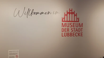 Museum der Stadt Lübbecke Logo und Schriftzug Willkommen im Museum der Stadt Lübbecke an einer weißen Wand.