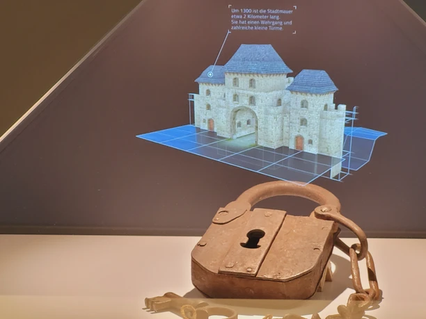 Schloss eines Stadttores vor digitaler 3D-Rekonstruktion des Tores und der Lübbecker Stadtmauer. Rostiges Vorhängeschloss mit Kette vor digitaler 3D-Rekonstruktion der Lübbecker Stadtmauer.