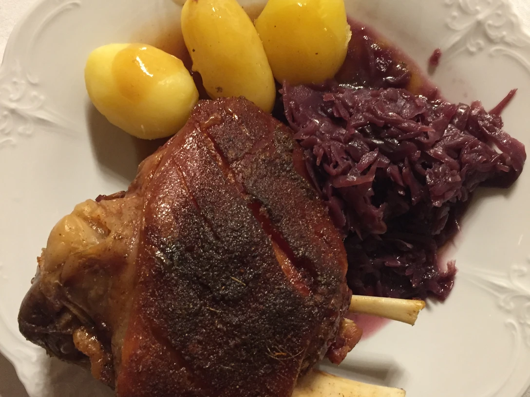 Gegrillte Schweinshaxe, serviert mit drei Salzkartoffeln und einer Portion Rotkohl auf einem weißen Teller.