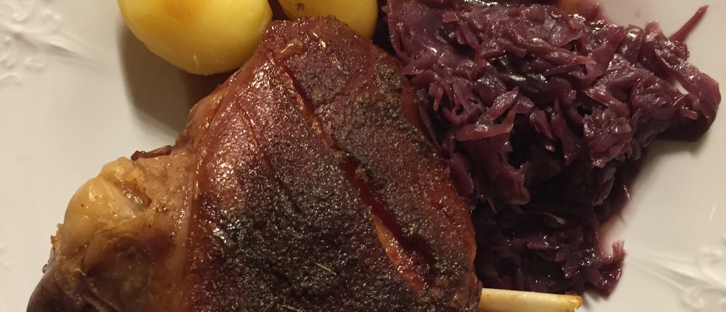 Grilled pork knuckle, served with three boiled potatoes and a portion of red cabbage on a white plate.